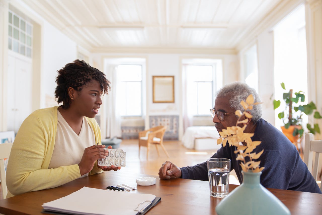 Home An elderly man sits with a caregiver discussing medication at a table in a bright room.